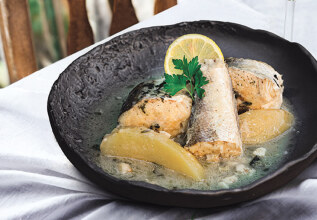 A plate of fish with lemon slices and herbs, accompanied by a glass of white wine, placed on a table with a striped cloth next to a wooden chair.