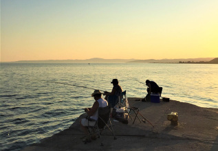 Three people fishing from a concrete pier at sunset with a calm sea and distant hills in the background.