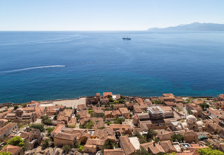 Aerial view of the historic coastal town of Monemvasia, Greece, with terracotta rooftops and the Mediterranean Sea in the background, featuring a distant oil rig.