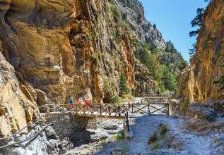 Scenic view of Samaria Gorge in Crete, featuring a wooden bridge crossing a rocky riverbed with visitors walking along the path, surrounded by steep cliff walls and lush greenery.