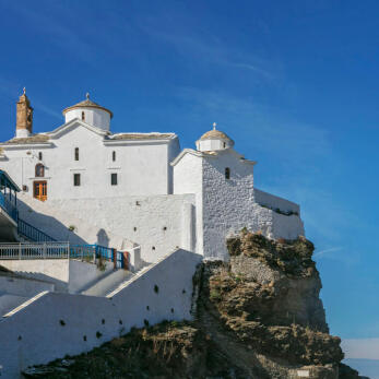 Large white church with a dome and stone roof with a smaller church and stairs on a rock above the sea. 