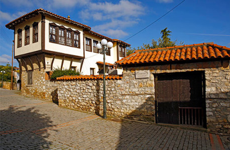 Paved road with stone fence and tiles. Large wooden front door. On the left there is a traditional two-storey building with tiles and a closed ledge with wooden windows.
