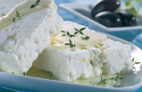 Δυο κομμάτια τυρί φέτα με λίγο λάδι και φρέσκια ρίγανη σε ένα άσπρο πιάτο και λίγες μαύρες ελιές σε ένα μικρότερο πιάτο.