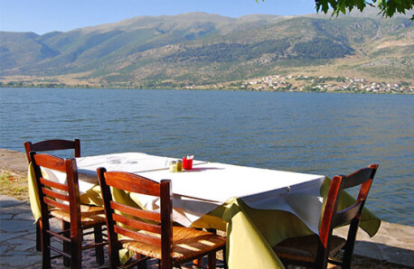 Τραπέζι με ξύλινες καρέκλες δίπλα σε λίμνη. Στο βάθος βουνά και παραλίμνιος οικισμός. Καλοκαίρι με λιακάδα.