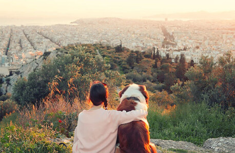 Woman with dog sitting and looking at the view of the city from a green hill.