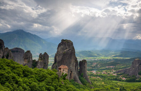 Row of high cliffs over a valley. In front a large monastery. In the back mountains and the sun's rays. 