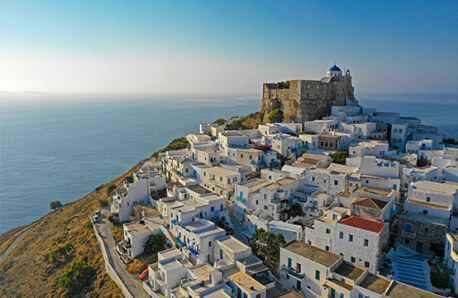 A serene Greek village with whitewashed buildings and a blue-domed church atop a hill, overlooking the Aegean Sea under a clear sky.