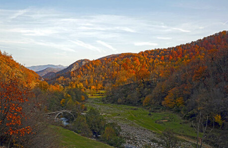 Landscape with green grass and river with stone arched bridge. All around hills with trees with brown and red leaves or no leaves. Blue sky with a few clouds.