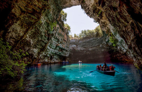 Cave with turquoise waters and an opening at the top where the sky and trees can be seen. Boats with people on the water. 