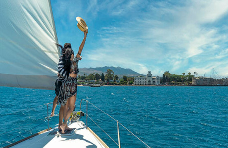 Sailing boat with a man and a woman standing in front. They salute towards land. Trees and buildings can be seen.