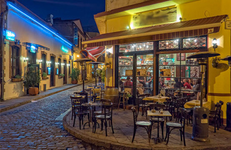 Cobblestone street with small, illuminated buildings. Tavern with tables outside and people inside. Night.