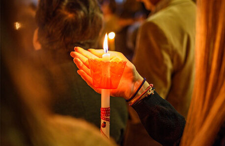 White lit candle with red protector. A hand protects the flame, among people. Night.