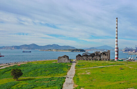 Aerial photograph of an open coastal area with low vegetation and an abandoned factory with a large chimney. In the background, sea, ships and islands. Sunshine and light clouds.