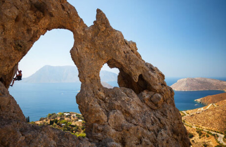Cliff with 2 large holes. Behind them you can see land, sea and islands. Man climbs inside the biggest hole. 
