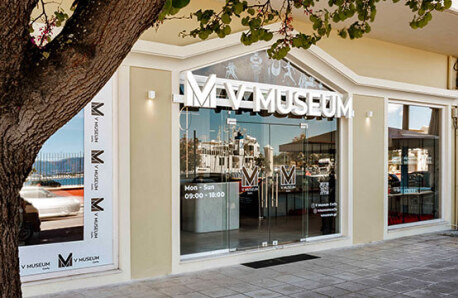 Glass museum facade with the museum's name, framed by a light-colored wall. Reflecting low buildings and a clear sky.