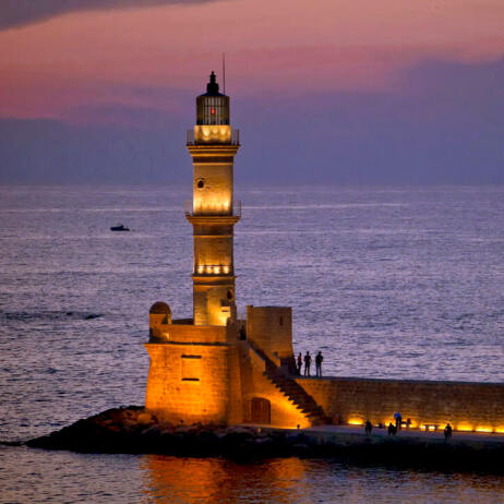 Old lighted lighthouse made of stone on the edge of a pier with a few people at sunset. 