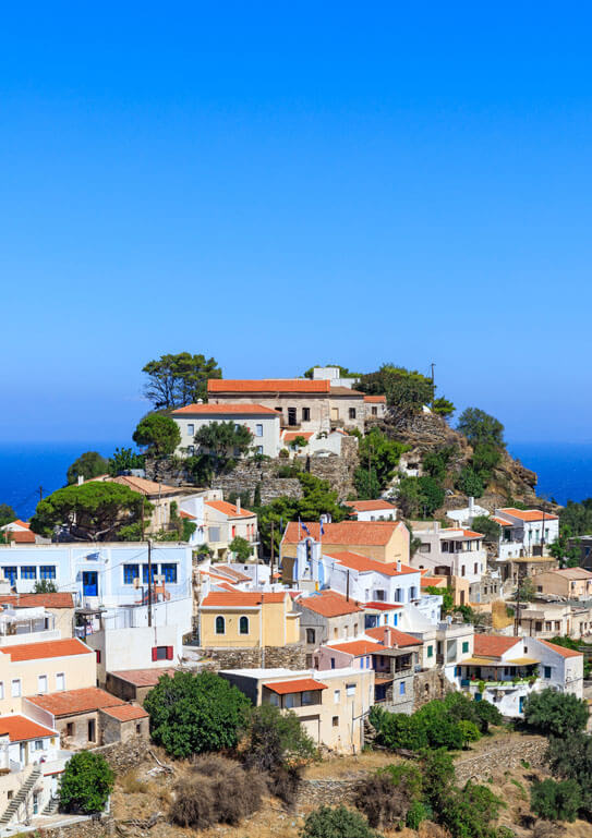 Picturesque settlement at the top of a hill. Tiled houses with some trees in between. In the background is the sea. 