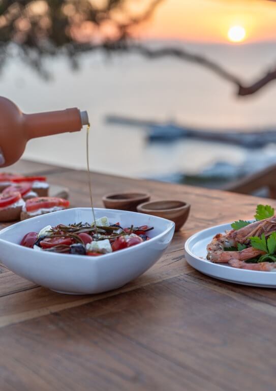 olive oil being served in a plate with greek salad, next to it there is dish with shrimps, and the sunset in the background