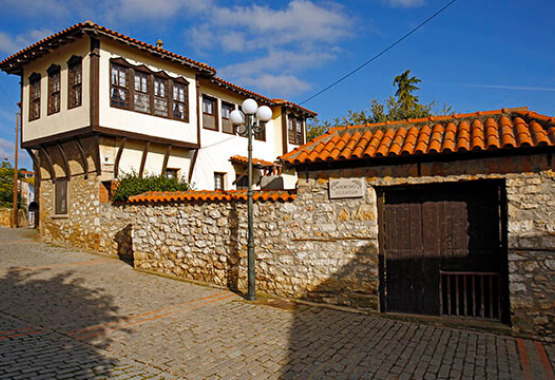 Paved road with stone fence and tiles. Large wooden front door. On the left there is a traditional two-storey building with tiles and a closed ledge with wooden windows.