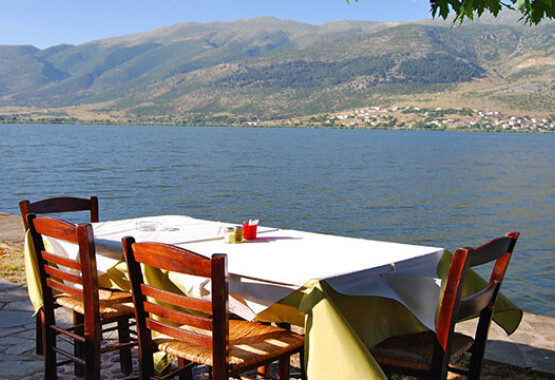 Table with wooden chairs by a lake. In the background, mountains and a lakeside settlement. Summer with sunshine.