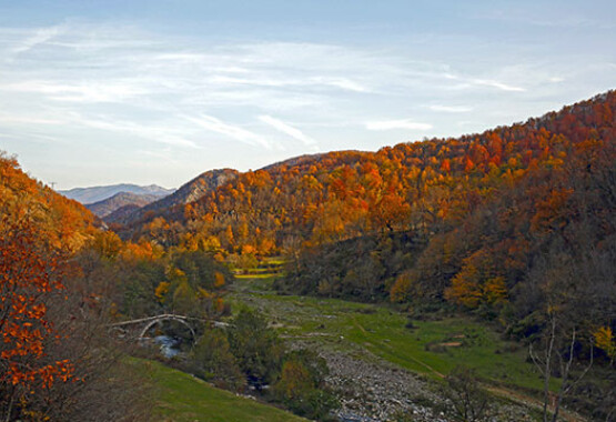 Landscape with green grass and river with stone arched bridge. All around hills with trees with brown and red leaves or no leaves. Blue sky with a few clouds.
