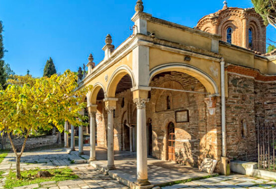 Byzantine stone church with dome and arches in the courtyard. Paved yard with small tree.