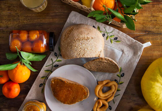 Wooden table with a basket of citrus fruits, rustic bread, plate with bread and jam, juice, jam and buns.