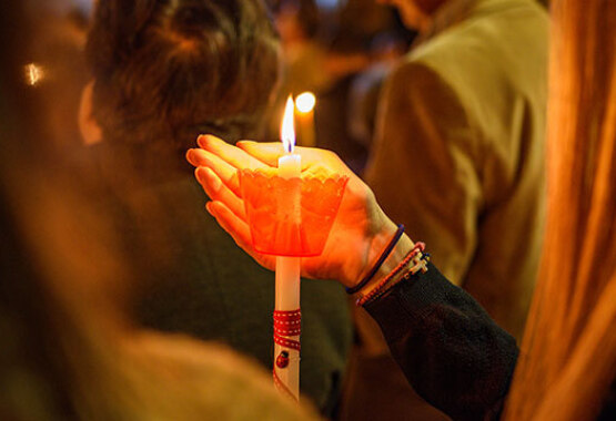 White lit candle with red protector. A hand protects the flame, among people. Night.