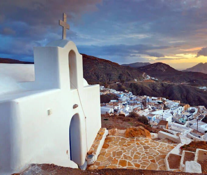 Μικρή άσπρη εκκλησία πάνω από γραφικό οικισμό στο βάθος σκοτεινοί λόφοι το ηλιοβασίλεμα. 