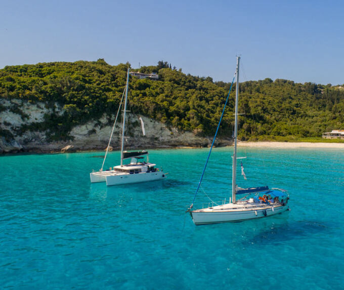 Παραλία του νησιού με γαλαζοπράσινα νερά, πεύκα και δύο ιστιοφόρα στο βάθος