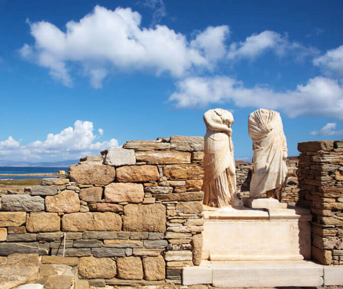 Archaeological site with low stone walls and 2 headless statues. In the background is the sea. 