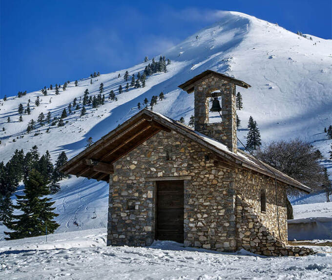 A small stone-built church with a snowy mountain behind in Kalavryta