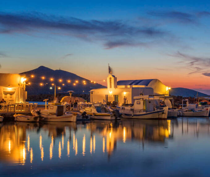 Peaceful harbor with boats and a small white church with lights at sunset. 