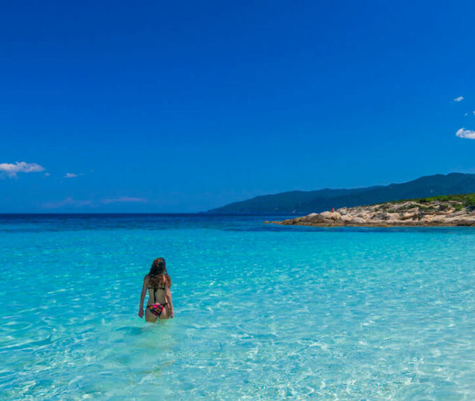 Shallow blue sea and a girl in a swimsuit enters. In the background mainland with low vegetation. 