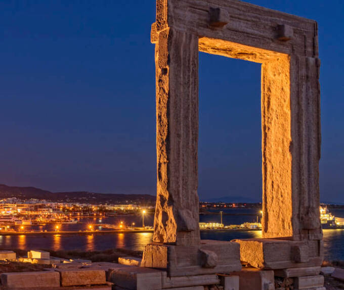 Night view of a large ancient gate in the foreground and in the background a Cycladic settlement with a port. 