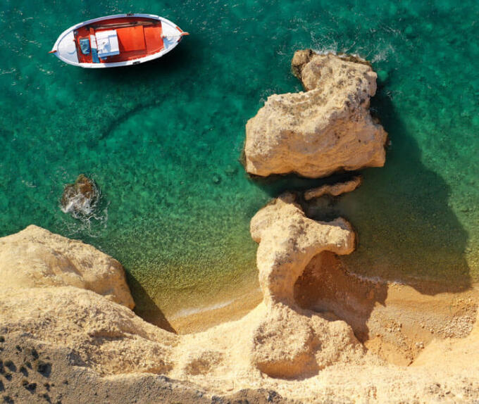 Aerial view of a white and red boat in crystal clear sea next to small beaches surrounded by rocks. 