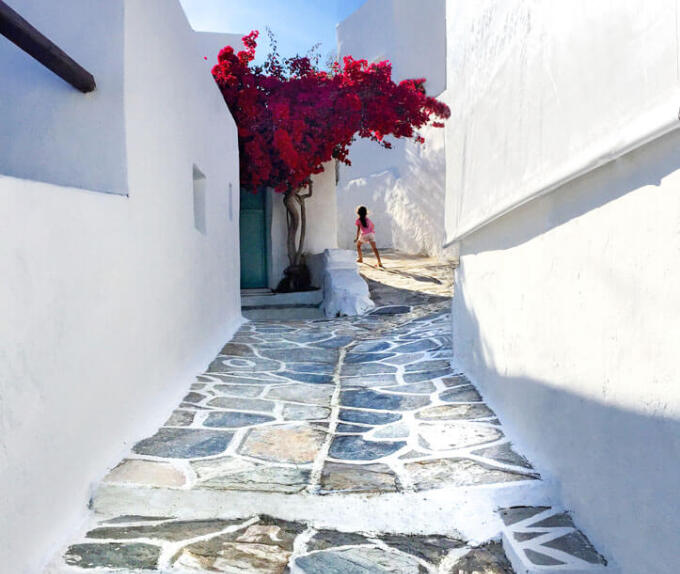 Girl walking on cobbled alley between white houses. In a yard a large red bougainvillea. 