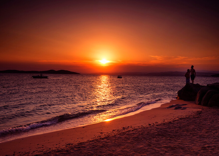 two persons standing on a beach at sunset, observing the horizon where the sun is setting over the ocean. Two boats float nearby, enhancing the tranquil scene.