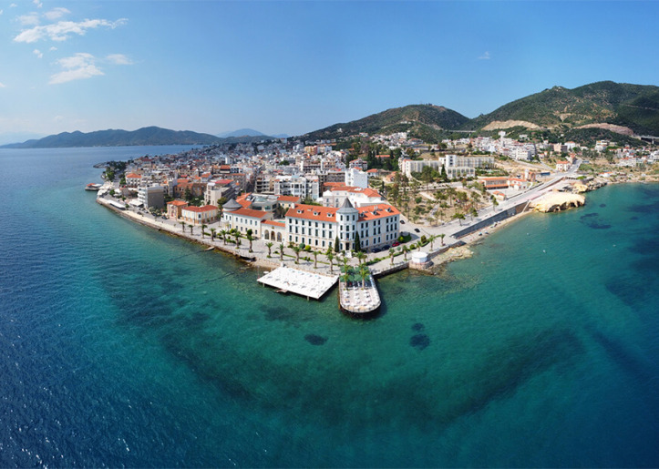 Aerial view of Cesme, a coastal town in Turkey, showing vibrant buildings, boats docked at a pier, and clear blue waters surrounded by hills.