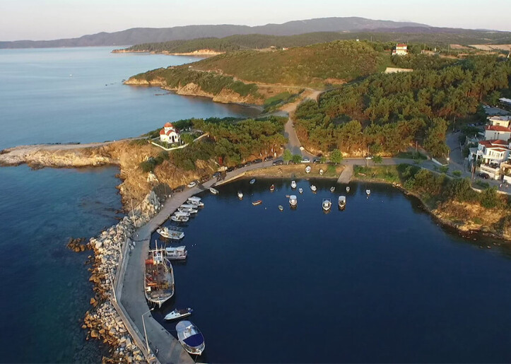 Aerial view of a small marina with several boats anchored, adjacent to a rocky coastline with scattered buildings, overlooking a calm sea and rolling hills in the background.