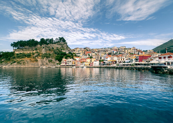 View of the picturesque coastal town of Parga, Greece, with colorful buildings cascading down the hillside, reflecting in the clear blue water of the Ionian Sea under a bright sky.