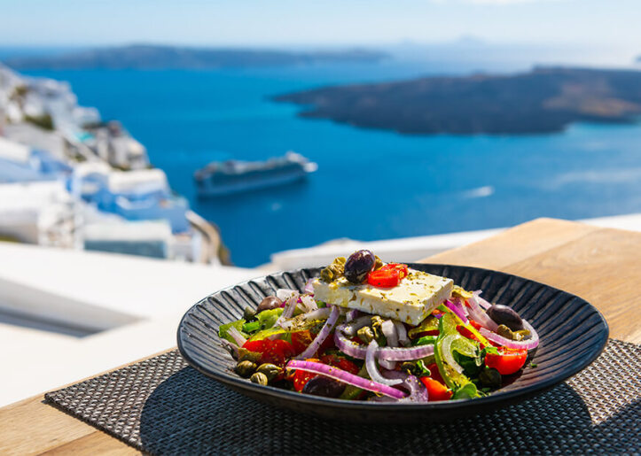 A plate of Greek salad with feta cheese, olives, tomatoes, and onions, served on a table overlooking the caldera in Santorini, with the deep blue sea and a ship in the background.