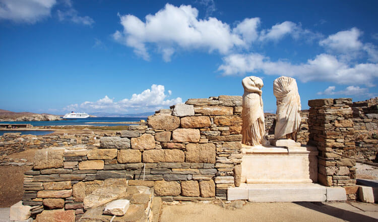 Archaeological site with low stone walls and 2 headless statues. In the background is the sea. 