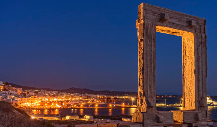 Νυχτερινή λήψη μεγάλης αρχαίας πύλης σε πρώτο πλάνο και στο βάθος κυκλαδίτικος οικισμός με λιμάνι.