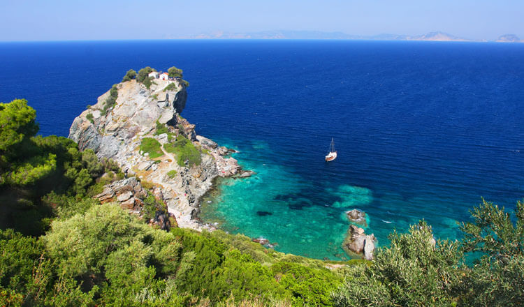 Ακτογραμμή με πράσινη βλάστηση και ένα μεγάλο βράχο στη θάλασσα. Στην κορυφή του μια μικρή εκκλησία. Στη θάλασσα ένα σκάφος.