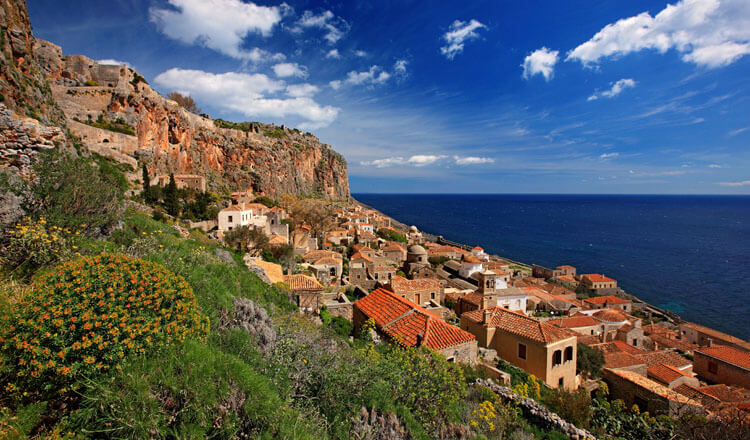 Traditional stone settlement on a hillside above the sea. High rock behind. Low vegetation in front. 