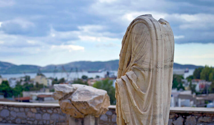 Άποψη της πόλης από ψηλά, με ένα αρχαίο άγαλμα χωρίς κεφάλι να ατενίζει την πόλη και το λιμάνι