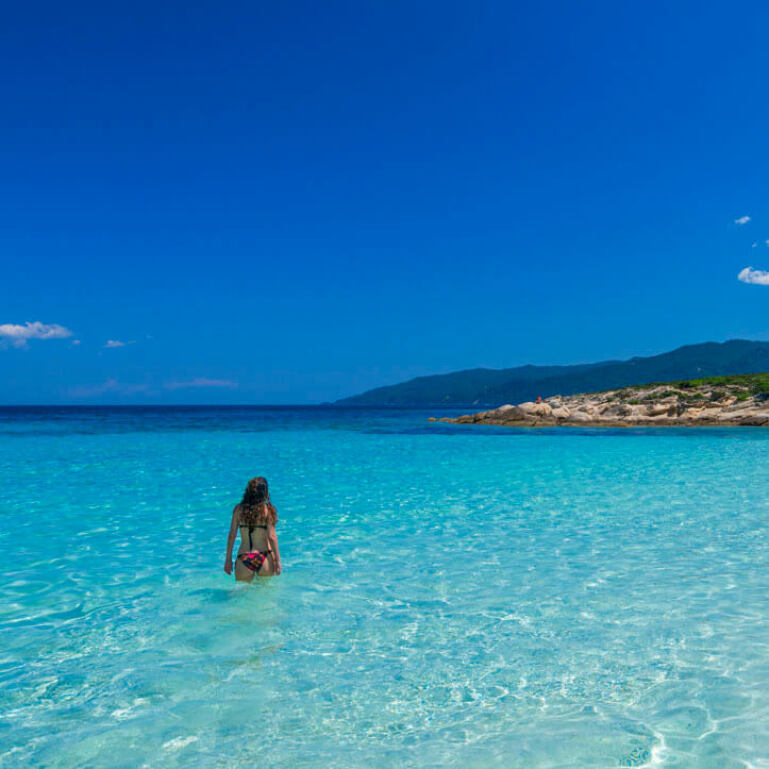 Shallow blue sea and a girl in a swimsuit enters. In the background mainland with low vegetation. 