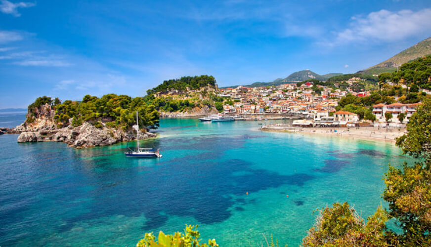 Sea with transparent waters, small green island, sailing boat, beach and in the background a picturesque settlement. 