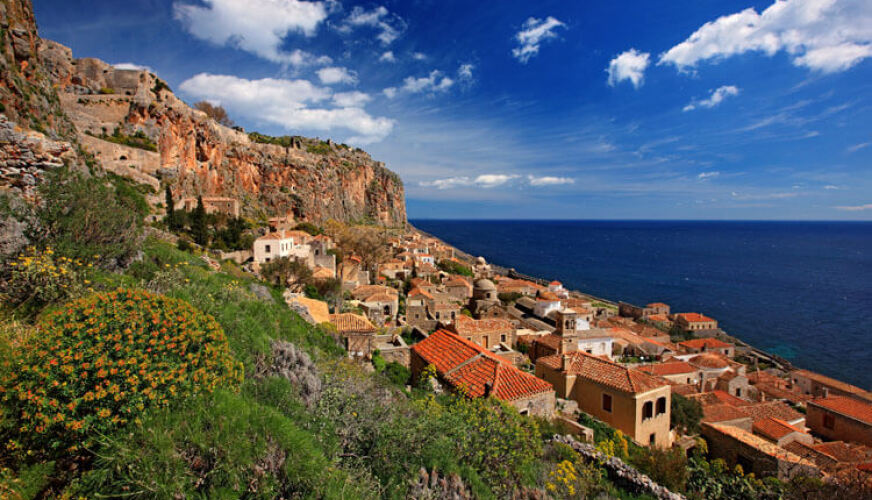 Παραδοσιακός πέτρινος οικισμός στην πλαγιά λόφου πάνω από τη θάλασσα. Ψηλός βράχος από πάνω. Χαμηλή βλάστηση μπροστά.